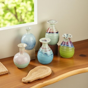Könnte beinhalten: Eine Sammlung kleiner, dekorativer Keramikvasen in verschiedenen Farben, darunter Hellblau, Rosa und Grün. Einige Vasen haben gestreifte Muster und gerüschte Ränder. Die Vasen stehen auf einer Holzoberfläche in der Nähe eines Fensters.