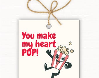May include: Square gift tag with the text "You make my heart POP!" in red. A cartoon popcorn box with a smiling face and arms and legs is also featured. The tag is cream-colored and has a jute string attached.