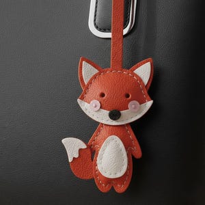 May include: A leather fox-shaped charm in orange, white, and black, hanging from a matching orange strap. The fox has white-tipped ears, pink cheeks, and a white belly. The charm is attached to a black bag.