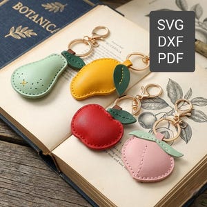 May include: A selection of fruit-shaped keyrings in green, yellow, red, and pink. Each keyring features a gold-coloured clasp and a small green leaf. The keyrings are arranged on an open book. The image also contains the text 'SVG DXF PDF'.