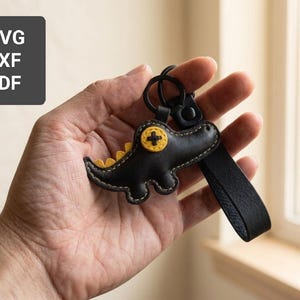 May include: Black leather keychain in the shape of a dinosaur with yellow accents. The keychain has a black strap and a metal ring. The image also includes the text "SVG, DXF, PDF".