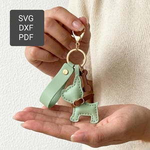 May include: A light green leather dog keyring with a brown collar and bone charm. The keyring has a gold-coloured ring and a matching green strap. The image also includes the text "SVG, DXF, PDF" in a dark grey box.
