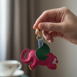Peut inclure: Un porte-clés éléphant en cuir rose avec des ailes vertes et des détails jaunes. Le porte-clés est attaché à un fermoir doré et une sangle bleue. L'éléphant a une trompe enroulée et une queue en forme de cœur.