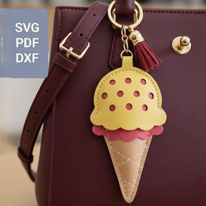 Peut inclure: Un porte-clés en forme de cornet de glace jaune et rose avec une chaîne et un pompon dorés. Le porte-clés est attaché à un sac à main bordeaux avec une sangle assortie. Le porte-clés porte le texte "SVG PDF DXF" dans le coin supérieur gauche.