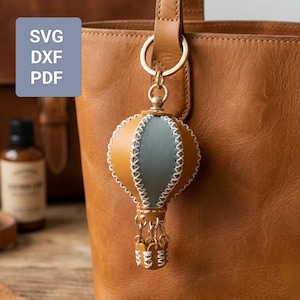 Puede incluir: Un llavero de globo aerostático de cuero marrón y gris con costuras blancas, sujeto a un bolso de cuero marrón. El llavero tiene un anillo dorado. También se ve un cuadrado azul con el texto "SVG, DXF, PDF".