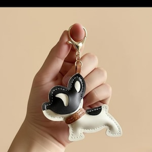 Può includere: Un portachiavi a forma di cane in pelle nera, bianca e marrone. Il portachiavi ha un fermaglio color oro e un collare marrone. Il design del cane presenta una testa nera e un corpo bianco con una macchia nera.
