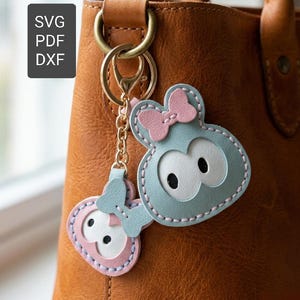 Könnte beinhalten: Zwei Schlüsselanhänger im Cartoon-Stil in Blau und Rosa mit einem Schleifendesign, die an einer goldfarbenen Kette und einem Verschluss befestigt sind. Sie hängen an einer braunen Lederhandtasche. Das Bild enthält auch den Text "SVG PDF DXF".