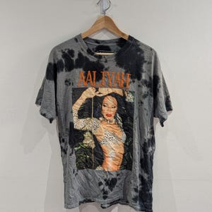 May include: A grey and black tie-dye t-shirt featuring a graphic print of Aaliyah in a leopard print top. The name AALIYAH is printed in orange letters above the image. The shirt has short sleeves, a crew neckline, and is displayed on a wooden hanger.