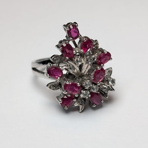May include: A silver-toned ring featuring a cluster of oval-cut deep pink rubies and small clear round-cut stones arranged in a floral-like pattern. The metal band is split at the sides, supporting the central decorative arrangement of gemstones.