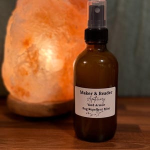 May include: A brown glass spray bottle with a black cap and a white label that reads "Maker & Reader Apothecary Yard Armor Bug Repellent Mist." An orange salt lamp is in the background.