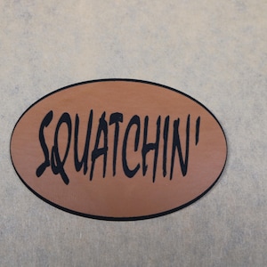 May include: An oval-shaped patch with a tan-colored surface and a thin black border. The word SQUATCHIN' is printed in the center in a bold, black, stylized font. The patch has a smooth, flat appearance and a distinct, dark outline.