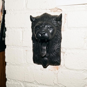 May include: A black lion head wall sculpture with an open mouth, mounted on a white brick wall. The sculpture has detailed fur and facial features. The lion's mouth is open, revealing a tongue. The sculpture is a decorative item.