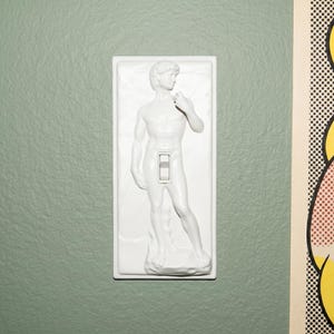 May include: White light switch cover featuring a sculpted relief of the statue of David. The switch is in the center of the cover, with the statue's form detailed in white. The cover is mounted on a light green wall.