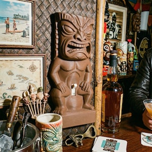 May include: A brown Tiki statue light switch cover with a carved face and muscular body. The switch is white. The background includes a framed map, a framed beach scene, and a bottle of liquor.