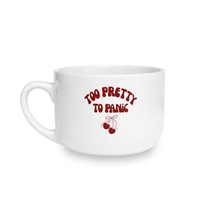 Puede incluir: Una taza de cerámica blanca con el texto en rojo oscuro "TOO PRETTY TO PANIC" arqueado sobre un gráfico de dos cerezas atadas con un lazo fino. La taza tiene un cuerpo redondeado y una sola asa curva en el lado izquierdo del recipiente.