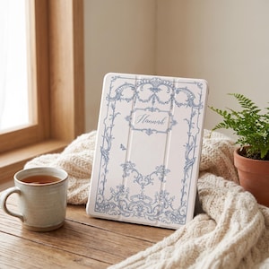May include: A white tablet case featuring a blue ornate scrollwork border and the name Hannah printed in a cursive script in the center. The case is standing upright, displaying a detailed decorative pattern with symmetrical floral elements.