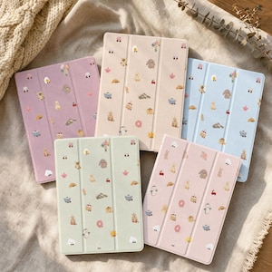 May include: Five rectangular tablet cases in pastel shades of purple, beige, light blue, sage green, and soft pink. Each case features a tri-fold design and is decorated with a repeating pattern of small, colorful icons including animals, food, and flowers.