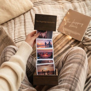 May include: A dark wooden box with an accordion-style photo strip extending upward. The top wooden panel features the text "The lingering memories." The photo strip displays four distinct outdoor scenes, including silhouettes of people at sunset.
