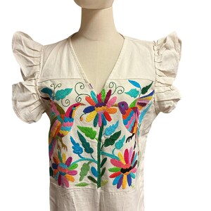 May include: An off-white blouse featuring ruffled sleeves and a V-neckline. The front panel displays vibrant, multi-coloured embroidery depicting two birds facing a central flower, surrounded by additional floral and leaf motifs in bright hues.