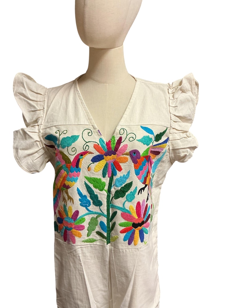 May include: An off-white blouse featuring ruffled sleeves and a V-neckline. The front panel displays vibrant, multi-coloured embroidery depicting two birds facing a central flower, surrounded by additional floral and leaf motifs in bright hues.