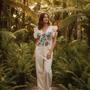 May include: An off-white jumpsuit featuring a V-neckline and ruffled sleeves. The bodice displays a vibrant, multi-coloured floral and bird embroidery pattern in shades of blue, red, green, and yellow. The garment has a fitted waist and wide-leg trousers.
