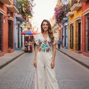 May include: A cream-coloured jumpsuit featuring a V-neckline and short ruffled sleeves. The bodice is decorated with vibrant, multi-coloured floral embroidery. The garment has wide-leg trousers and is paired with simple brown flat sandals.