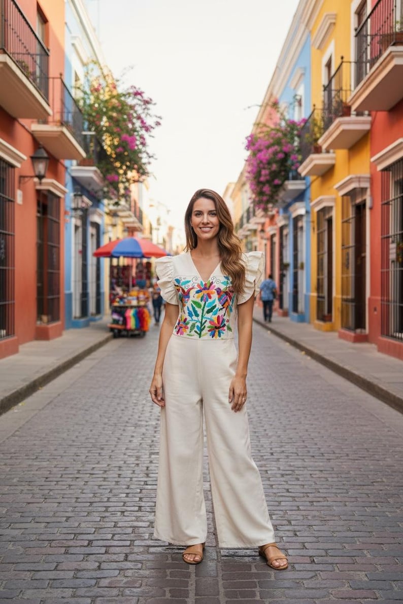 May include: A cream-coloured jumpsuit featuring a V-neckline and short ruffled sleeves. The bodice is decorated with vibrant, multi-coloured floral embroidery. The garment has wide-leg trousers and is paired with simple brown flat sandals.
