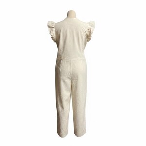 May include: An off-white jumpsuit featuring short flutter sleeves with ruffled edges, a round neckline, and a fitted bodice that transitions into straight-leg trousers. The garment is displayed on a mannequin, showcasing a clean, solid-coloured design.