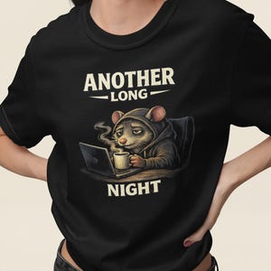 Op de afbeelding: Zwart t-shirt met een cartoonrat met een hoodie, zittend achter een laptop en een koffiemok vasthoudend. De tekst "ANOTHER LONG NIGHT" staat boven en onder de illustratie. De rat ziet er moe uit.