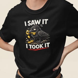 Puede incluir: Camiseta negra con un diseño de un cuervo sosteniendo una llave y monedas. El texto "I SAW IT" está encima del cuervo, y "I TOOK IT" debajo. El cuervo es negro.