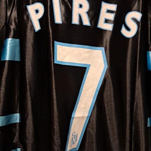 Op de afbeelding: Zwart shirt met de naam "PIRES" in witte letters met een lichtblauwe omlijning. Het nummer "7" is ook in wit met een lichtblauwe omlijning. Lichtblauwe strepen bevinden zich op de schouders en zijkanten.