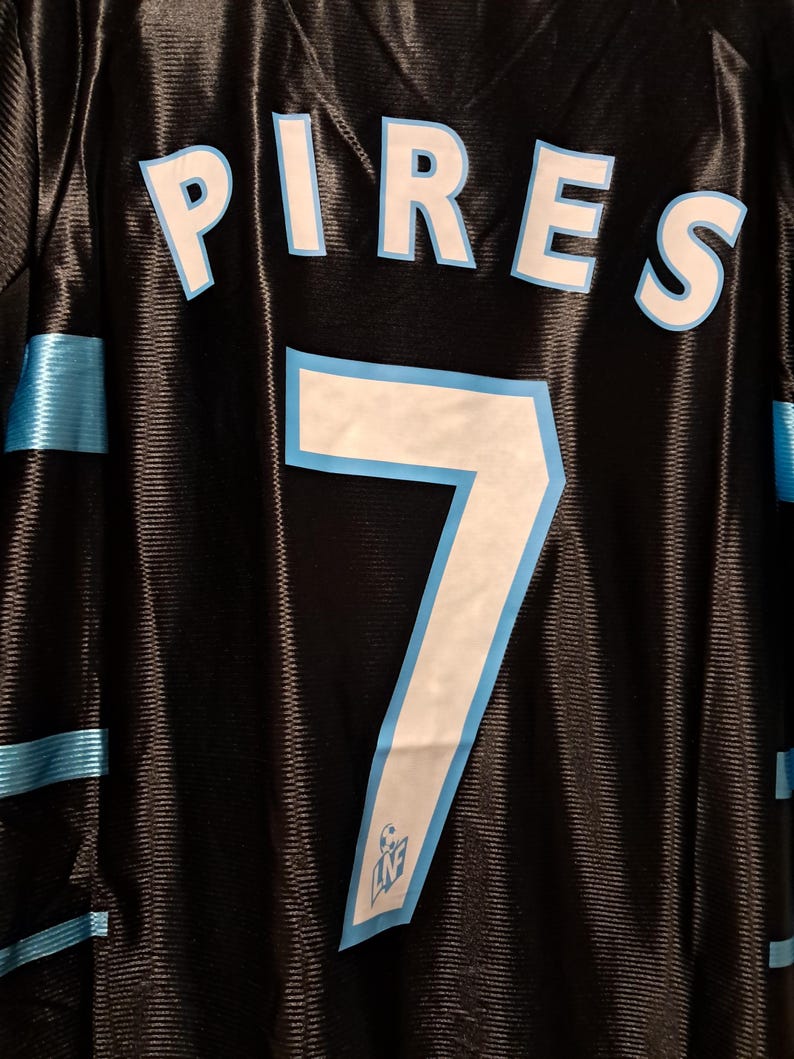 Op de afbeelding: Zwart shirt met de naam "PIRES" in witte letters met een lichtblauwe omlijning. Het nummer "7" is ook in wit met een lichtblauwe omlijning. Lichtblauwe strepen bevinden zich op de schouders en zijkanten.