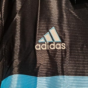 Op de afbeelding: Close-up van een zwart en blauw sportshirt. Het Adidas-logo is geborduurd in wit en lichtblauw. Het shirt heeft een geribbelde textuur en een horizontale blauwe streep aan de onderkant.
