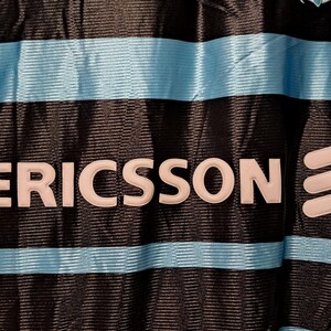 Op de afbeelding: Close-up van een zwart shirt met lichtblauwe horizontale strepen. Het woord "ERICSSON" staat in het wit afgebeeld, samen met een wit logo aan de rechterkant. Het shirt heeft een textuurpatroon.