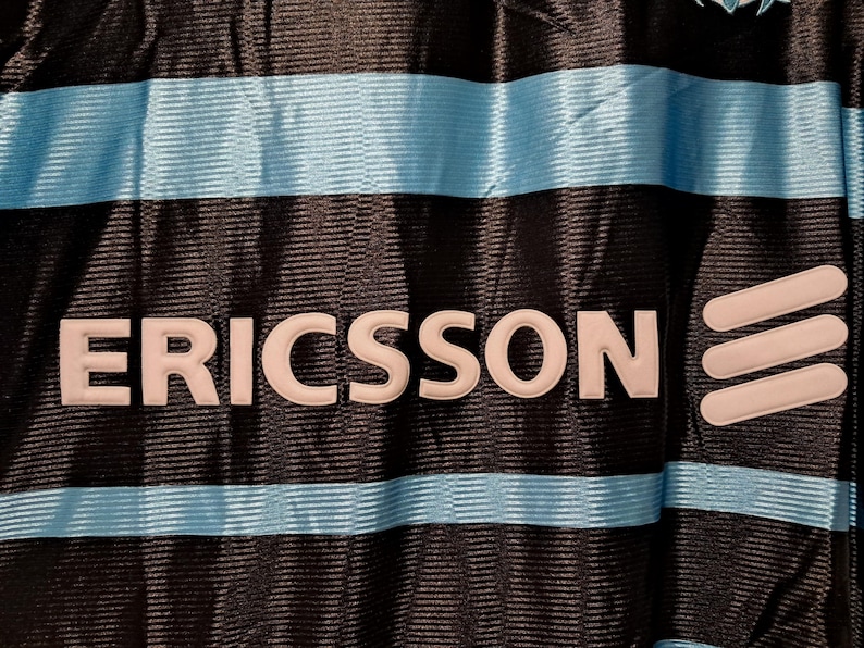 Op de afbeelding: Close-up van een zwart shirt met lichtblauwe horizontale strepen. Het woord "ERICSSON" staat in het wit afgebeeld, samen met een wit logo aan de rechterkant. Het shirt heeft een textuurpatroon.