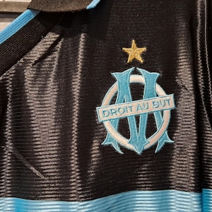 Op de afbeelding: Zwart shirt met lichtblauwe en zwarte kraag en bies. Het shirt heeft een lichtblauw en wit embleem met de tekst "DROIT AU BUT" en een gouden ster erboven. Het shirt heeft een geribbelde textuur.