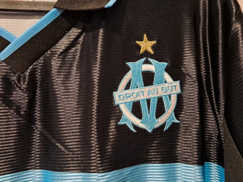 Op de afbeelding: Zwart shirt met lichtblauwe en zwarte kraag en bies. Het shirt heeft een lichtblauw en wit embleem met de tekst "DROIT AU BUT" en een gouden ster erboven. Het shirt heeft een geribbelde textuur.