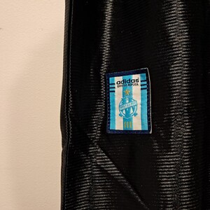 Op de afbeelding: Zwart sportshirt met een textuurpatroon. Een rechthoekige patch met het Adidas-logo en de tekst "OFFICIAL REPLICA" is bevestigd. De patch heeft een lichtblauw en wit ontwerp met een wapen.