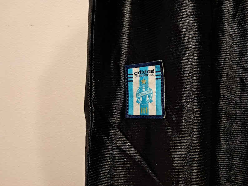 Op de afbeelding: Zwart sportshirt met een textuurpatroon. Een rechthoekige patch met het Adidas-logo en de tekst "OFFICIAL REPLICA" is bevestigd. De patch heeft een lichtblauw en wit ontwerp met een wapen.