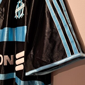 Op de afbeelding: Zwart en lichtblauw voetbalshirt met het Adidas-logo en het teamembleem. Het shirt heeft lichtblauwe strepen over de borst en op de mouwen. Het teamembleem is een lichtblauw wapen met de woorden "Olympique de Marseille".
