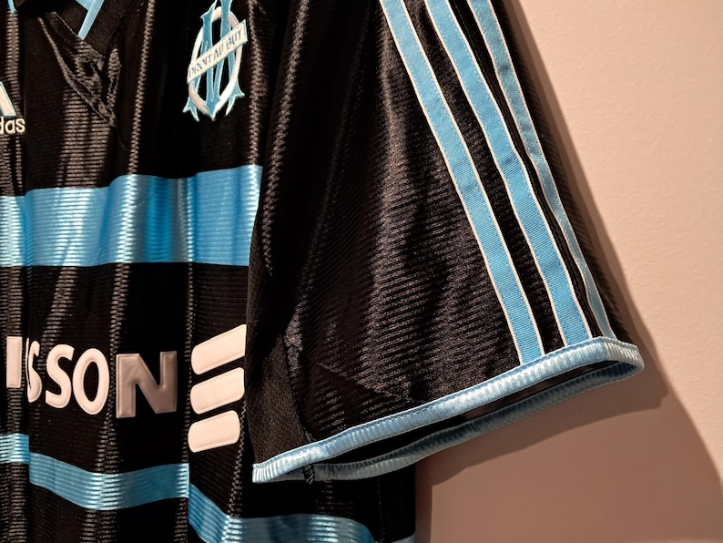 Op de afbeelding: Zwart en lichtblauw voetbalshirt met het Adidas-logo en het teamembleem. Het shirt heeft lichtblauwe strepen over de borst en op de mouwen. Het teamembleem is een lichtblauw wapen met de woorden "Olympique de Marseille".