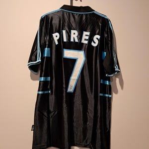 Op de afbeelding: Zwart voetbalshirt met de naam "PIRES" en het nummer "7" in wit en lichtblauw. Het shirt heeft lichtblauwe strepen op de mouwen en zijkanten. Het shirt hangt aan een zwarte hanger.