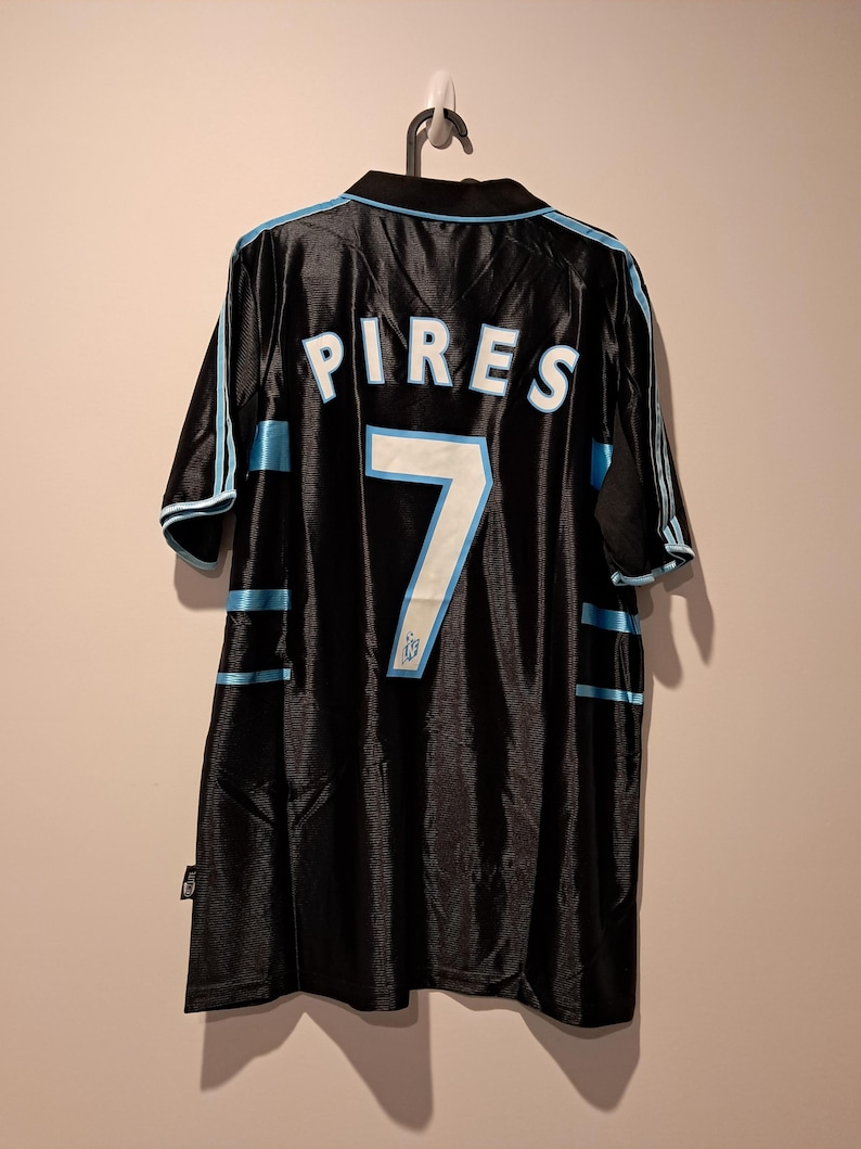 Op de afbeelding: Zwart voetbalshirt met de naam "PIRES" en het nummer "7" in wit en lichtblauw. Het shirt heeft lichtblauwe strepen op de mouwen en zijkanten. Het shirt hangt aan een zwarte hanger.