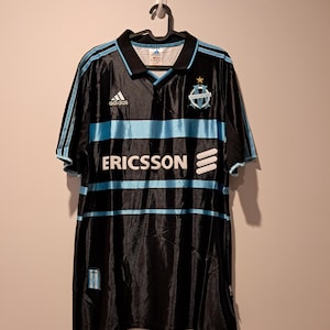 Op de afbeelding: Zwart voetbalshirt met lichtblauwe strepen en accenten. Het shirt heeft het Adidas-logo, het woord "ERICSSON" en een teamembleem. Het hangt aan een zwarte hanger tegen een neutrale achtergrond.