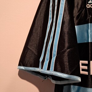 Op de afbeelding: Zwart voetbalshirt met lichtblauwe strepen en accenten. Het Adidas-logo is zichtbaar. Het shirt heeft het woord "ERIC" in witte letters. Lichtblauwe strepen lopen over de mouwen.
