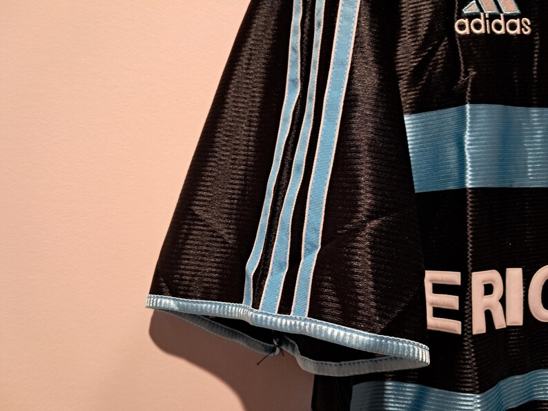 Op de afbeelding: Zwart voetbalshirt met lichtblauwe strepen en accenten. Het Adidas-logo is zichtbaar. Het shirt heeft het woord "ERIC" in witte letters. Lichtblauwe strepen lopen over de mouwen.