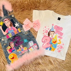 May include: A denim overall with pink feather trim and a white t-shirt. The overall has cartoon character appliques and the name "Aubrie". The t-shirt says "Can You Say 2" with cartoon characters.