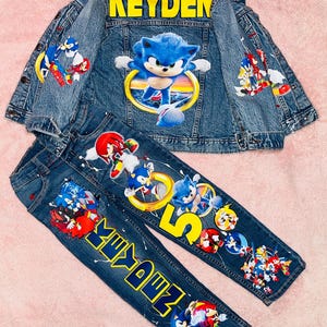 May include: A denim jacket and jeans set featuring Sonic the Hedgehog characters. The jacket has "KEYDEN" in yellow lettering on the back, with Sonic graphics. The jeans also have Sonic graphics and the name "KEYDEN".