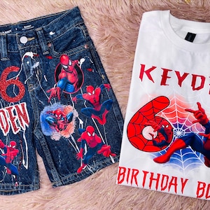 May include: A pair of dark blue denim shorts and a white t-shirt featuring Spider-Man graphics. The shorts have a red glitter number 6, the name KEYDEN, and white paint splatters. The t-shirt displays the name KEYDEN, a red number 6, and BIRTHDAY BOY.