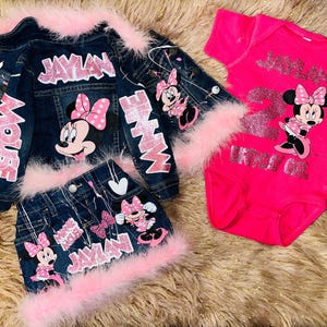May include: A denim jacket, skirt, and onesie set featuring Minnie Mouse graphics. The jacket and skirt have pink feather trim and the name "JAYLAN" in glitter. The onesie is hot pink with a Minnie Mouse graphic and the name "JAYLAN".